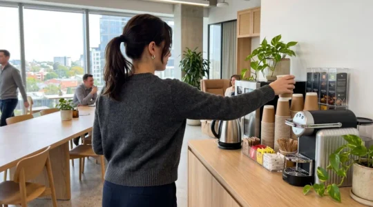 Un employé de bureau vu de dos tend la main vers un gobelet en carton posé sur un distributeur de boissons dans un espace pause lumineux et moderne